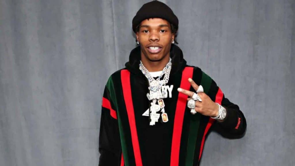 Lil Baby posing wearing black outfit with jewelry and hooded jacket.