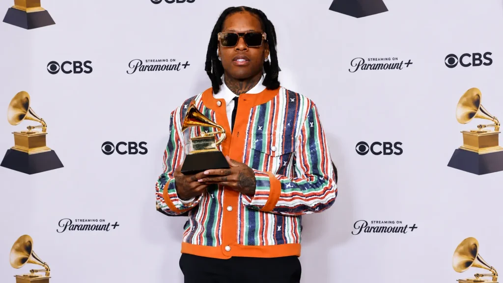 Lil durk holding a Grammy award while wearing a colorful striped jacket and sunglasses at a press event backdrop