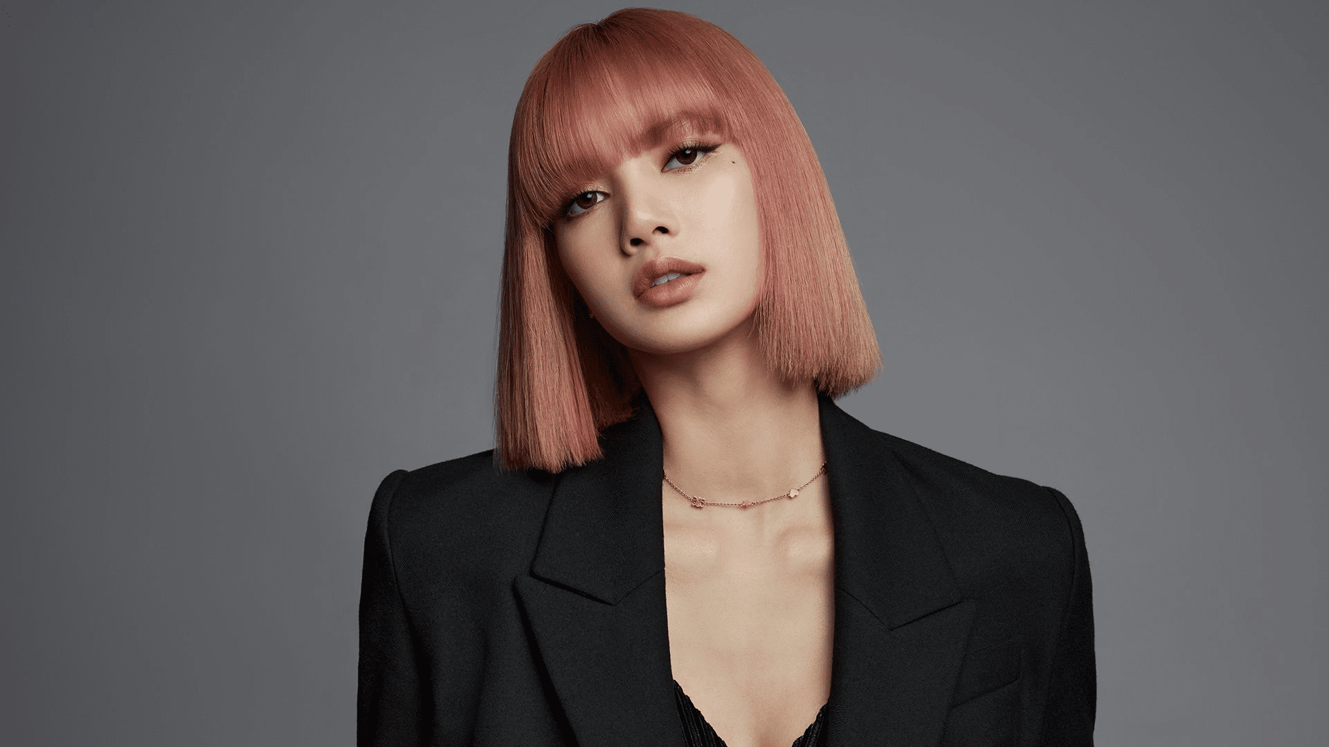 Lisa posing in elegant black blazer with pink hairstyle and neutral makeup against a smooth gray backdrop.