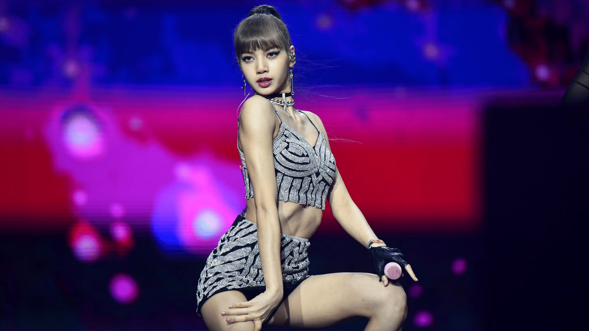 Lisa BLACKPINK performing on stage wearing a silver outfit with bright lights and colorful background during a live concert show