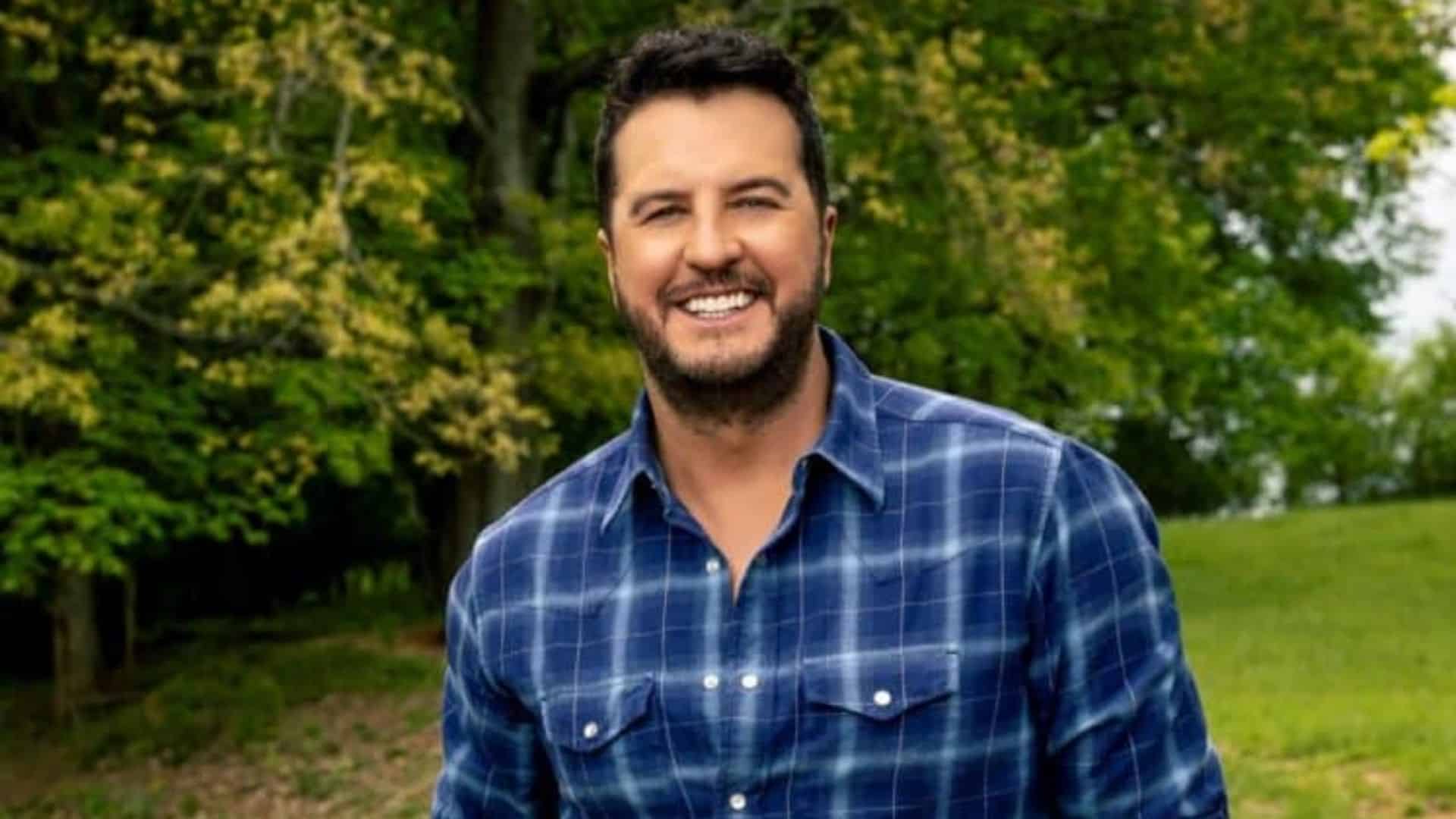 Luke Bryan smiles outdoors in a blue plaid shirt while standing on green grass with leafy trees filling the background