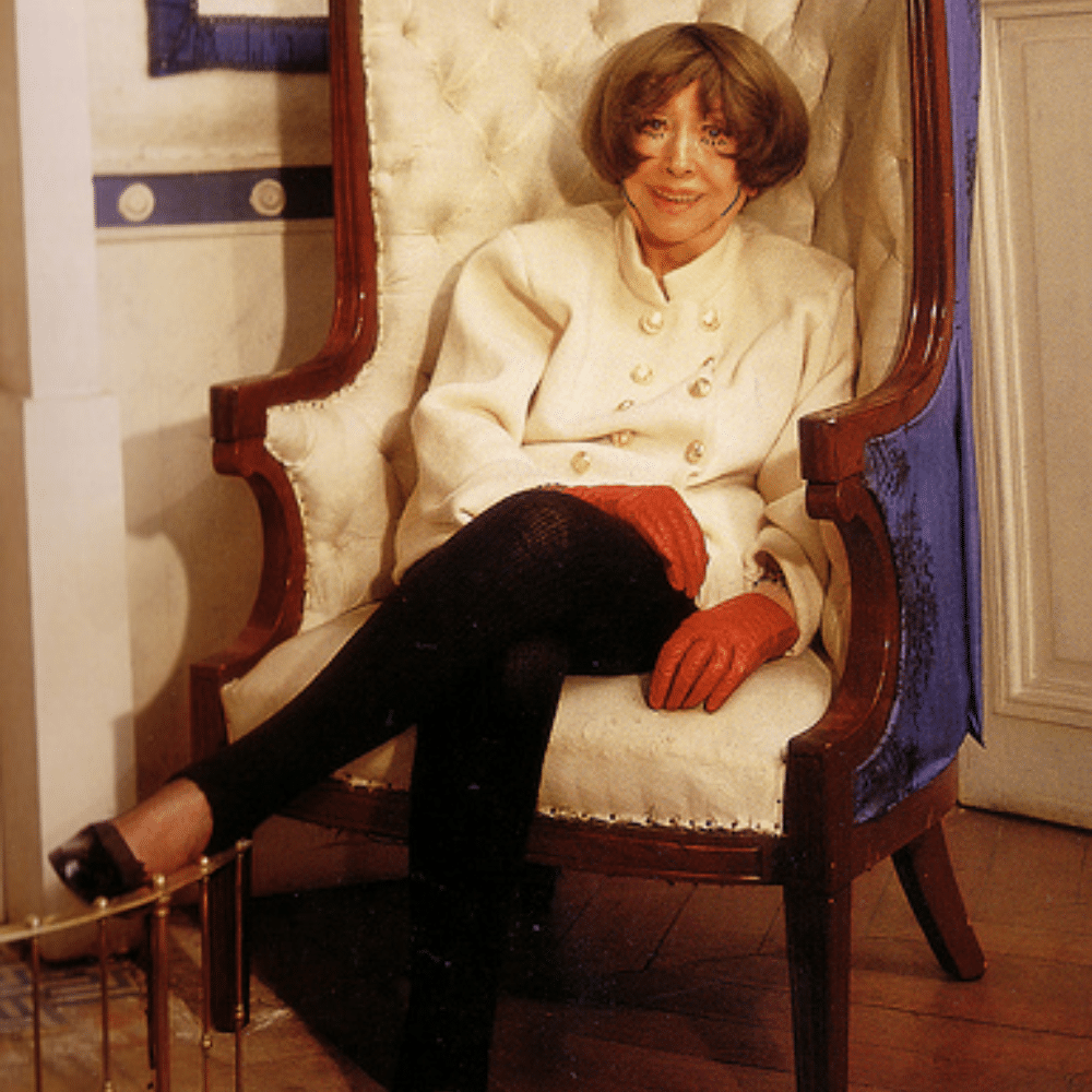 Madeleine Castaing in white coat and red gloves seated in tufted armchair in elegant interior with wooden floors.
