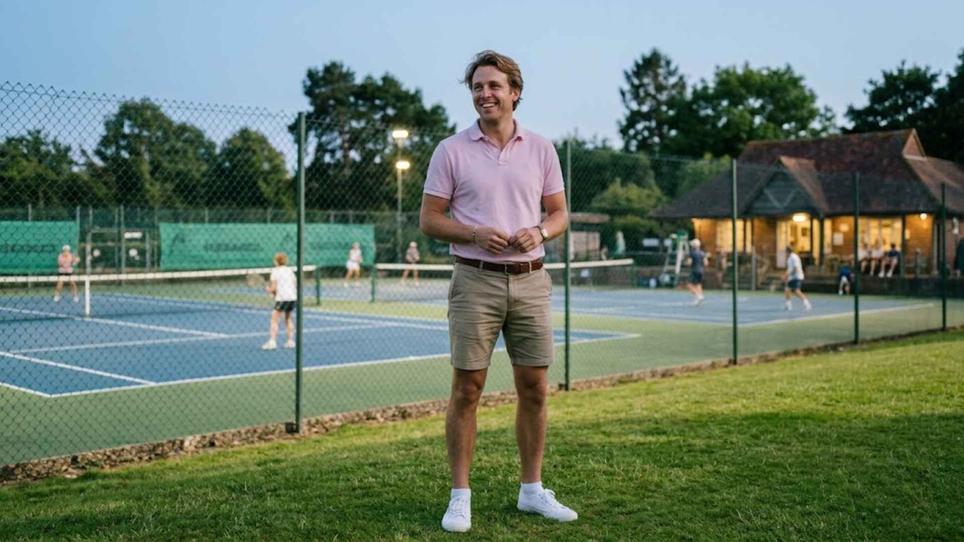 Man in a pastel pink polo shirt and khaki shorts standing on a lawn near a tennis court