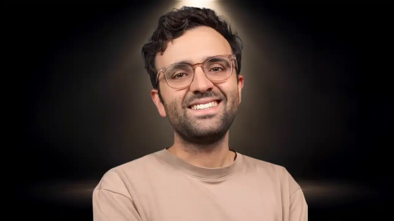 Man wearing clear-framed glasses smiling in a beige crewneck sweater against a dark backdrop