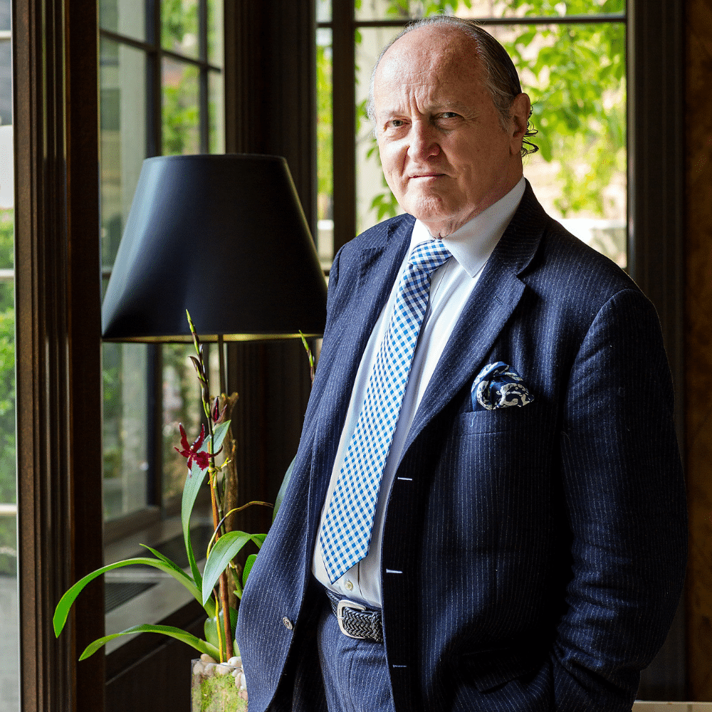 Mario Buatta in navy suit and patterned tie standing beside window and lamp in elegant office interior.