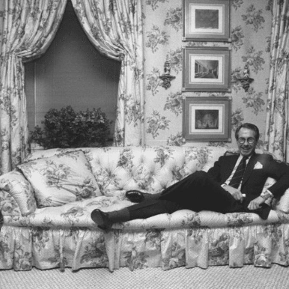 Black-and-white portrait of Mark Hampton reclining on floral sofa in richly patterned room with framed artwork and draped windows.