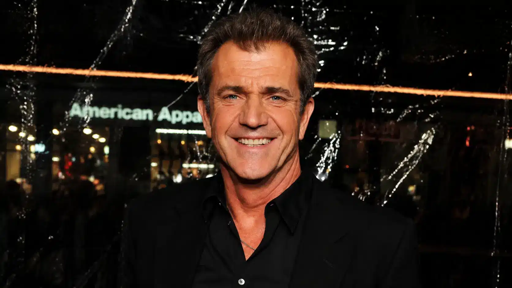 Mel Gibson in black suit posing at a nighttime event with lights and reflections creating a cinematic portrait background