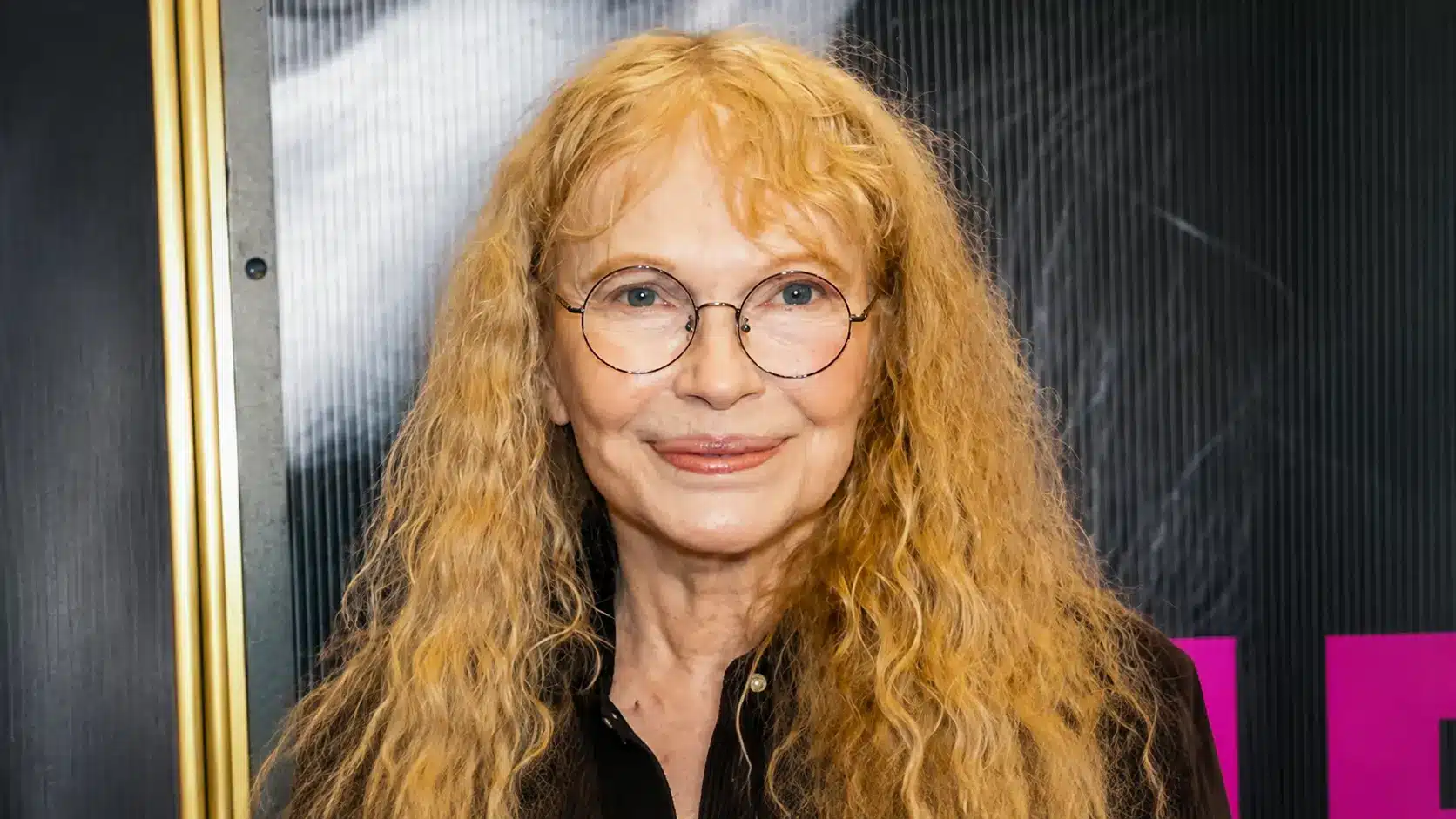 Mia Farrow with long blonde hair and round glasses smiling in a formal portrait with a dark event backdrop