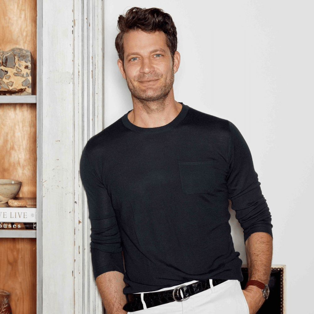 Nate Berkus in dark sweater and white pants leaning against wall beside wooden shelves in modern interior.