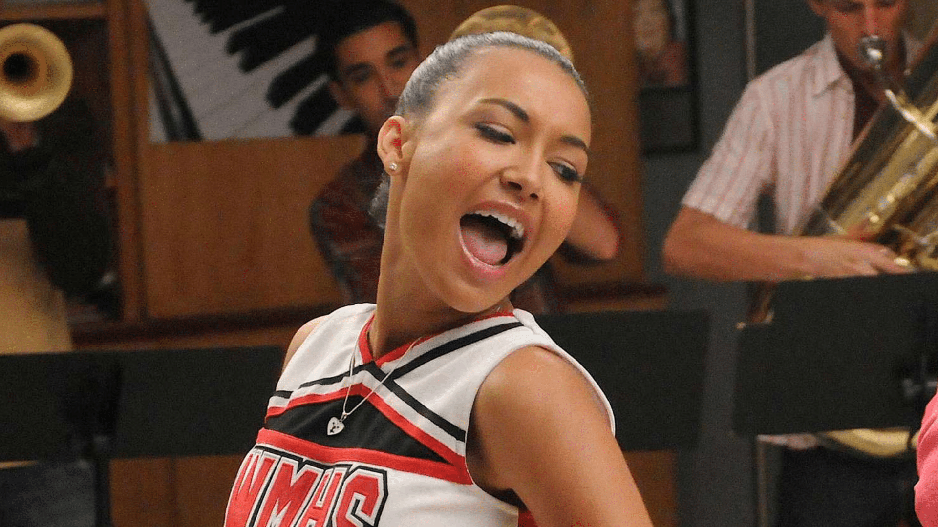 Naya Rivera performing as Santana Lopez in Glee wearing a cheerleading uniform during a musical scene with band members