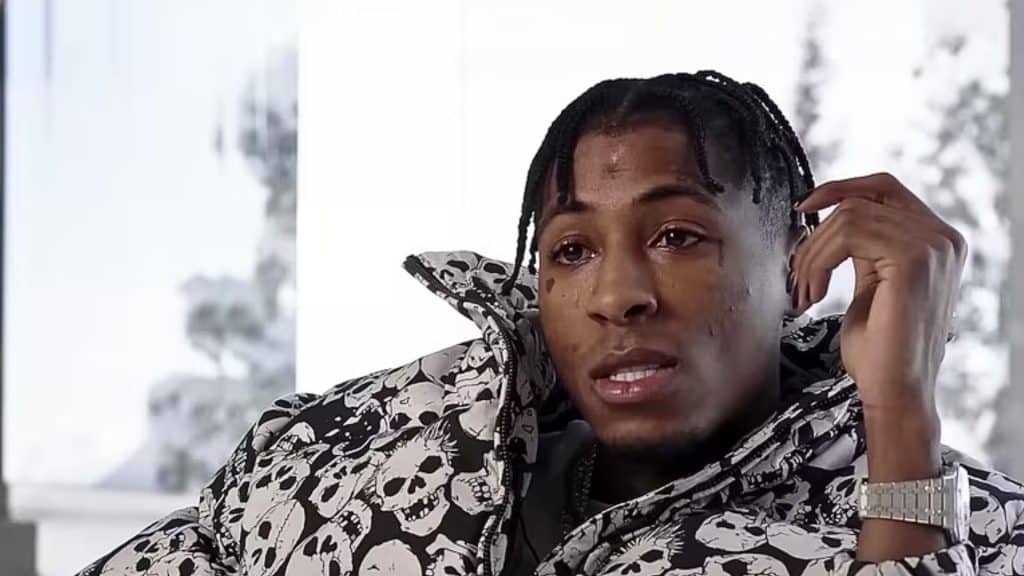 Nba youngboy with braided hair and sitting indoors in a skull printed jacket, looking toward the camera.