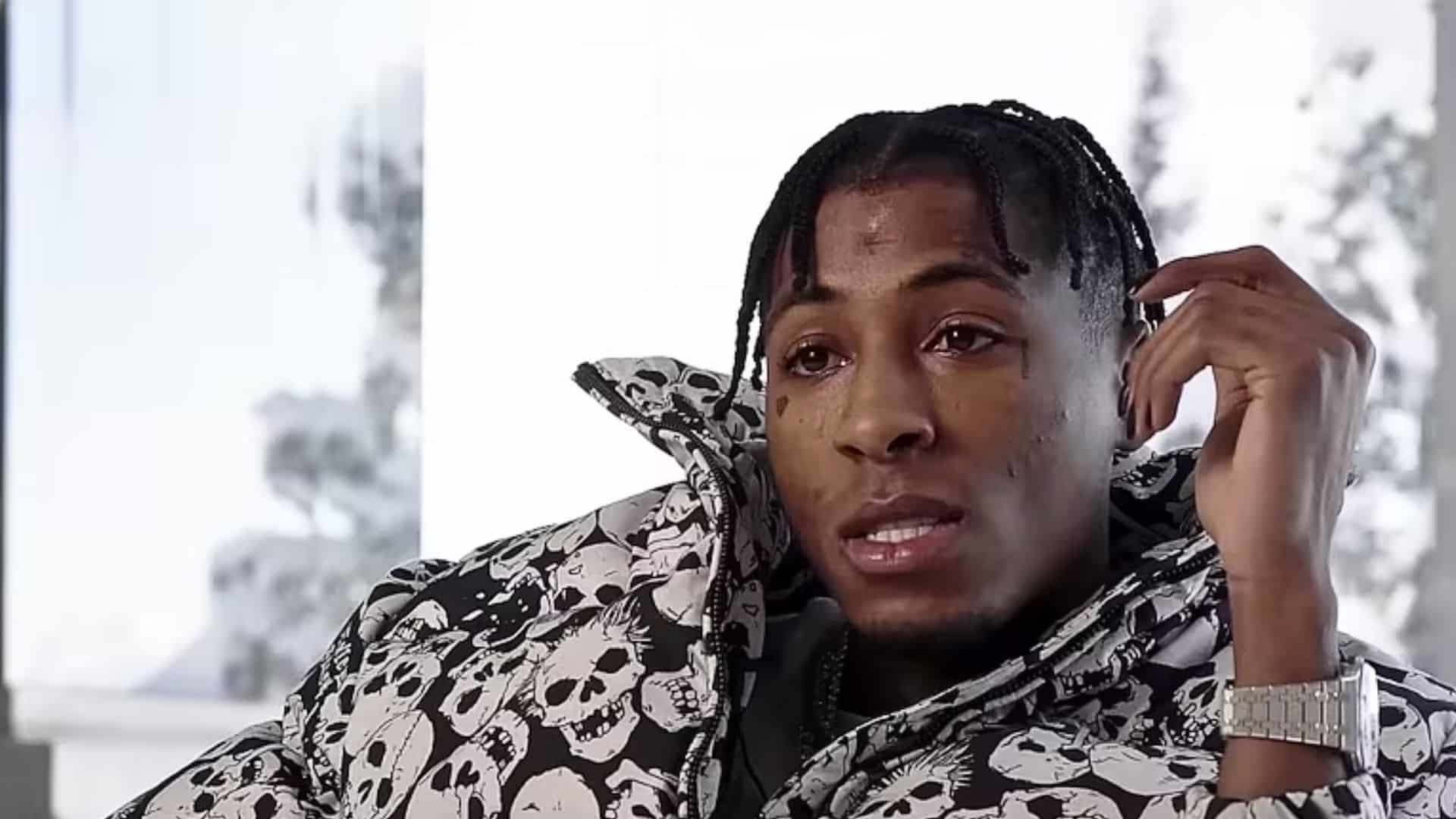 Nba youngboy with braided hair and sitting indoors in a skull printed jacket, looking toward the camera.