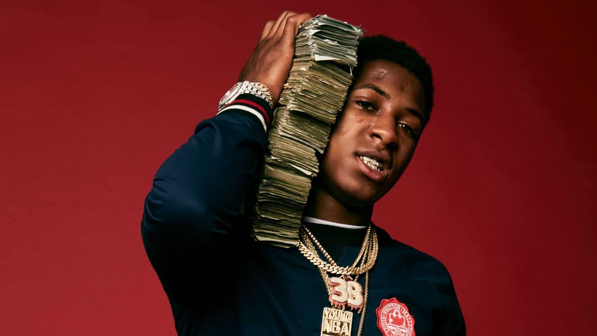 NBA YoungBoy posing against a red background with a large stack of cash, gold chains, a watch, and grills.