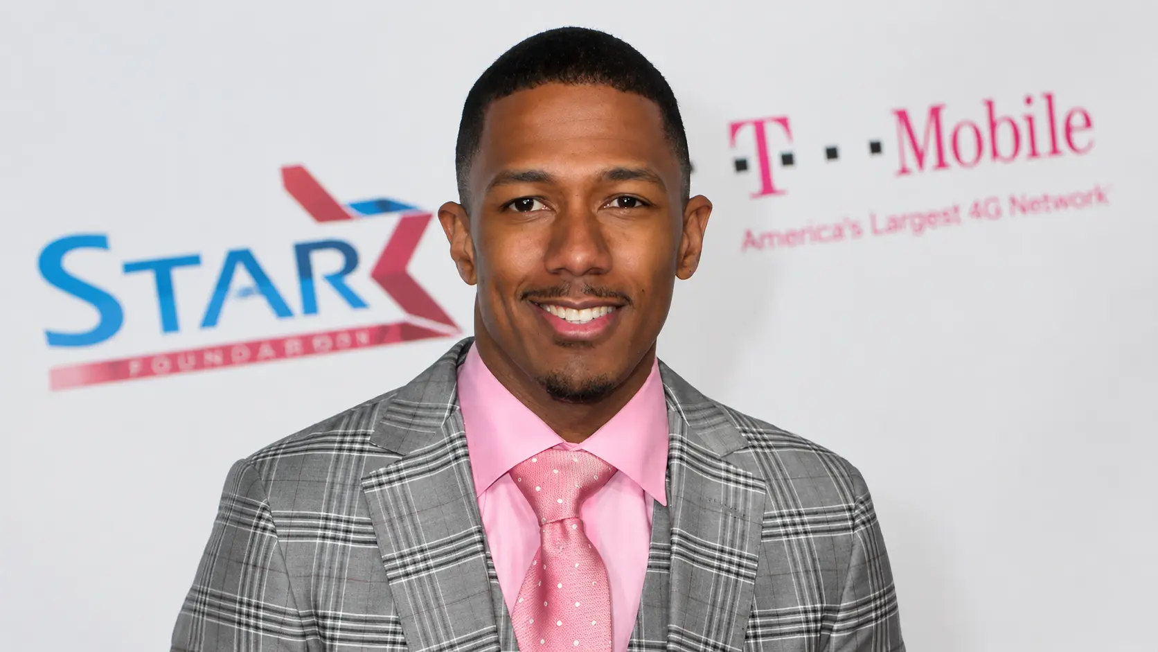 Nick Cannon wearing a gray plaid suit and pink shirt posing at an event with a branded white backdrop behind him