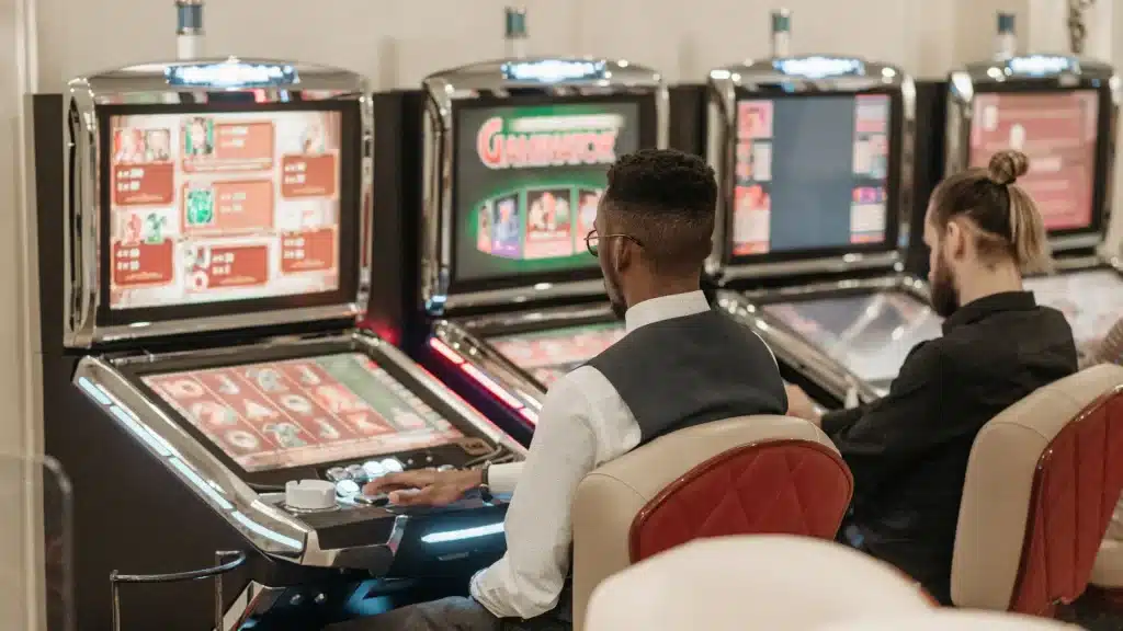 Two men seated at slot machines in a casino environment with soft lighting
