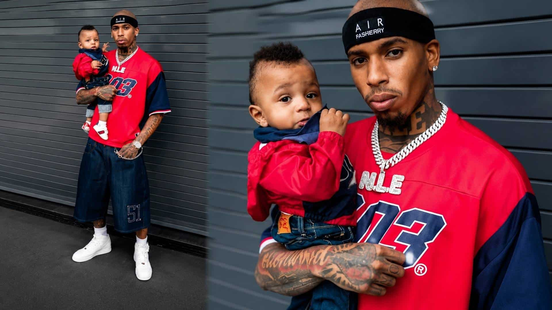 NLE Choppa holding his son ChoZen while posing in a red jersey and streetwear outfit.