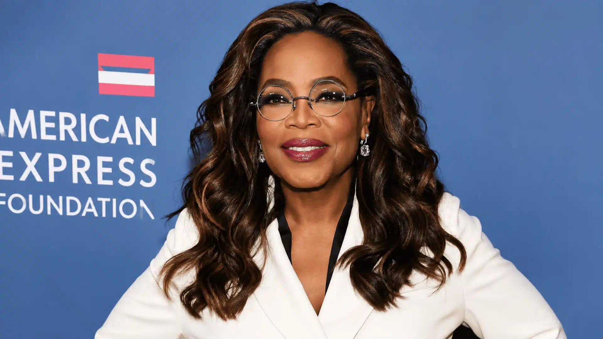 Oprah Winfrey wearing round glasses and a white blazer while posing against a blue event backdrop