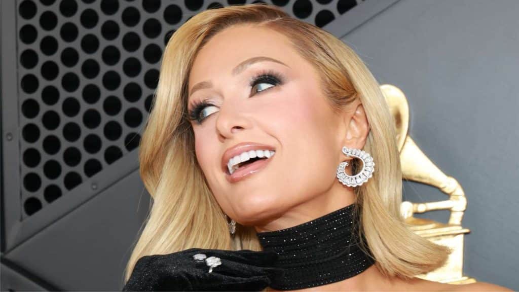 Paris Hilton smiling at a red carpet event wearing a black outfit with sparkling earrings and glamorous makeup