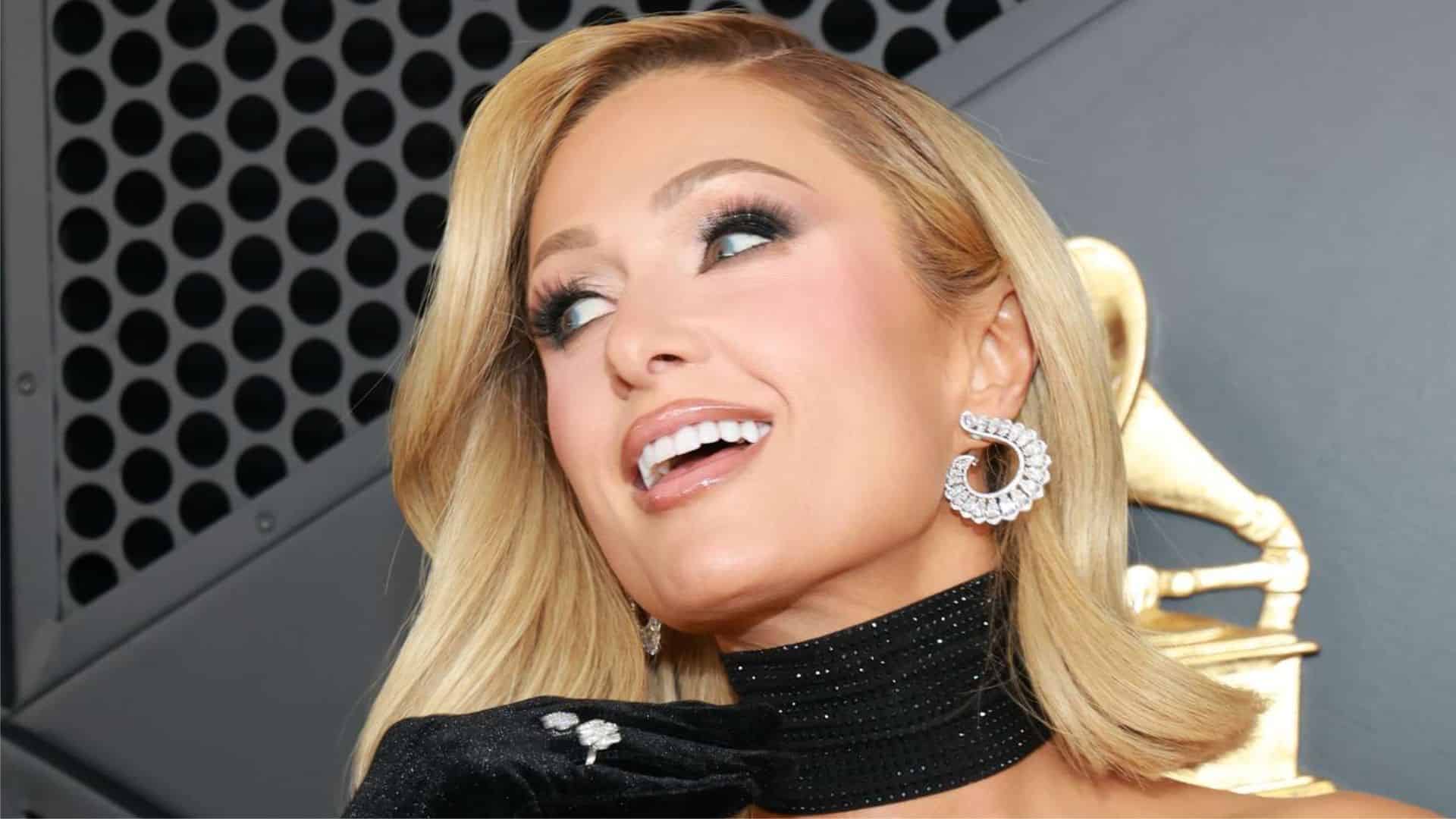 Paris Hilton smiling at a red carpet event wearing a black outfit with sparkling earrings and glamorous makeup
