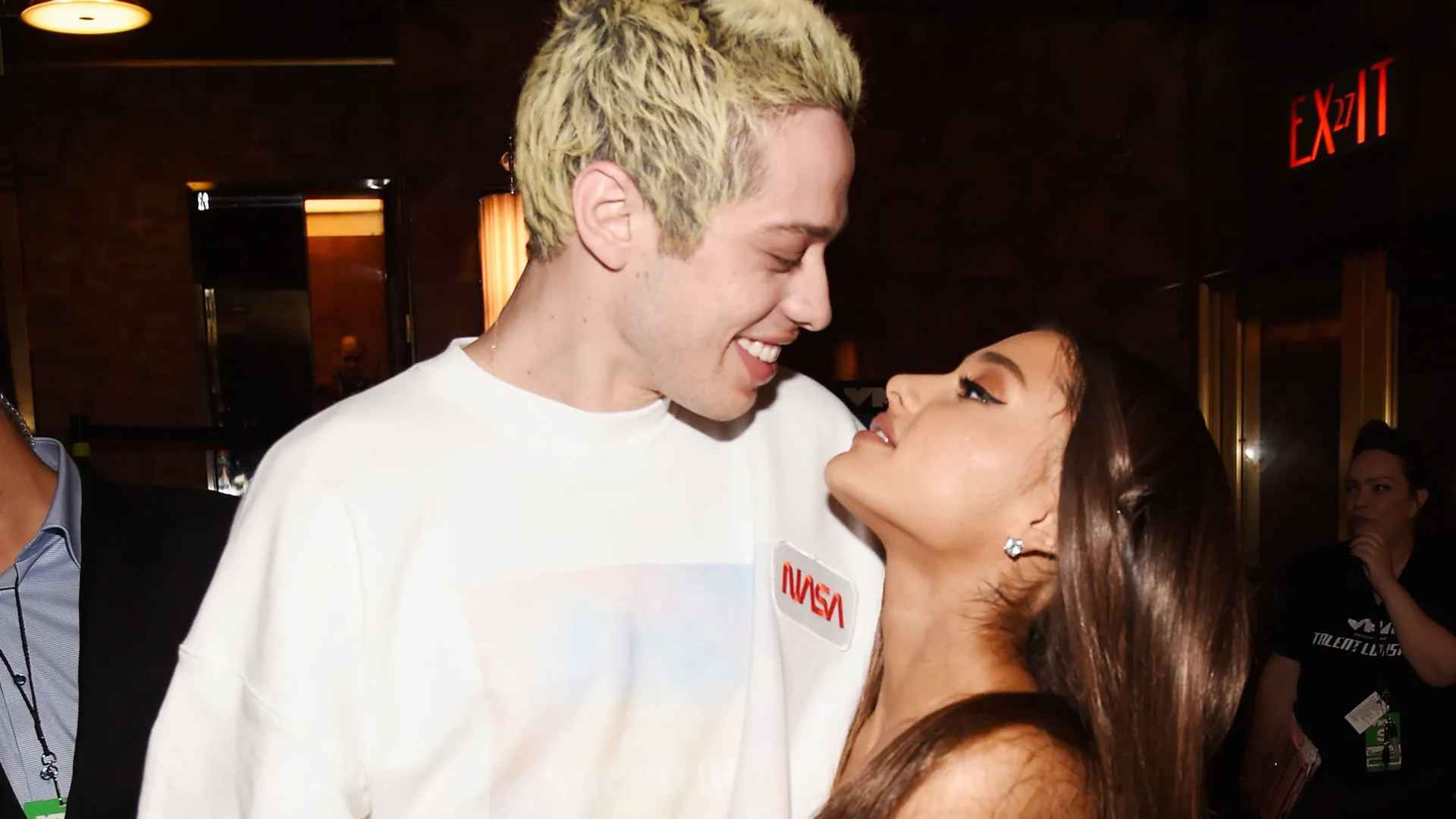 Pete andd Ariana standing close together at an indoor event with one person smiling while looking at the other under warm lights