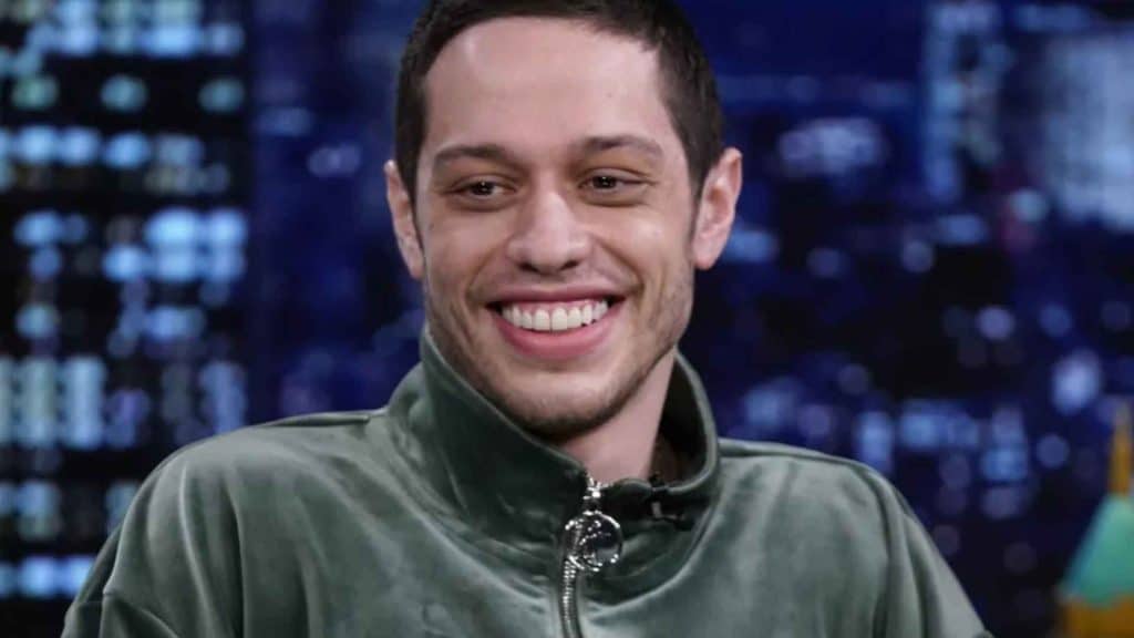 Smiling Pete Davidson wearing a green jacket during a studio appearance with a blurred blue background and bright lights behind him