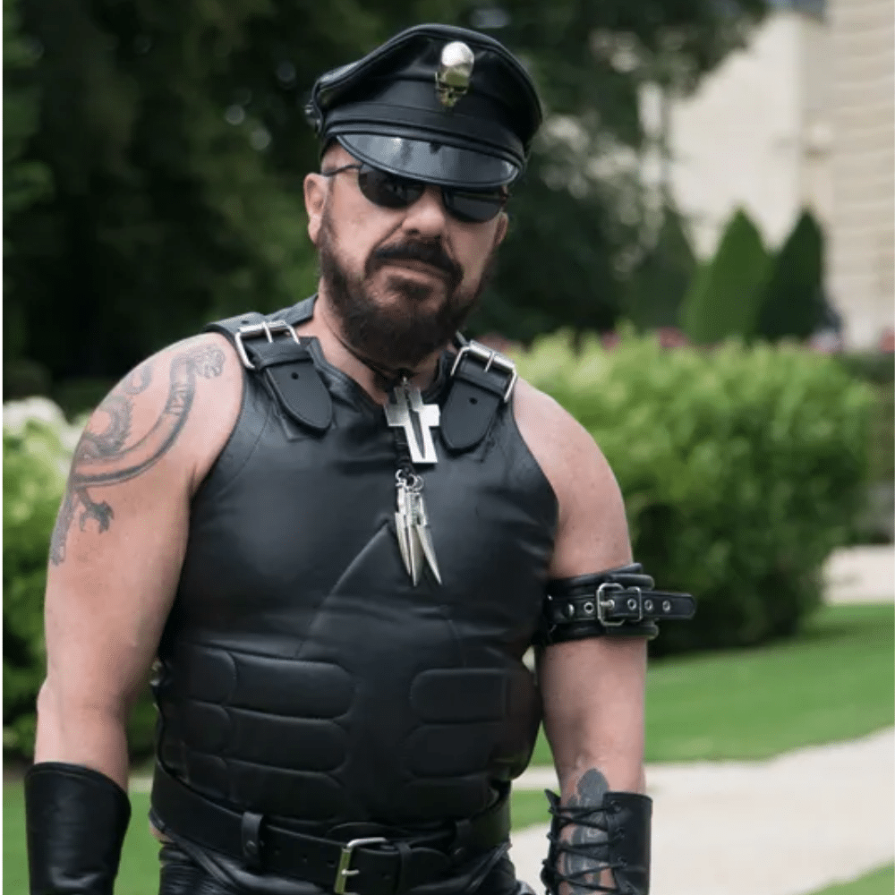 Peter Marino in black leather outfit, cap, and sunglasses standing outdoors on grassy path.
