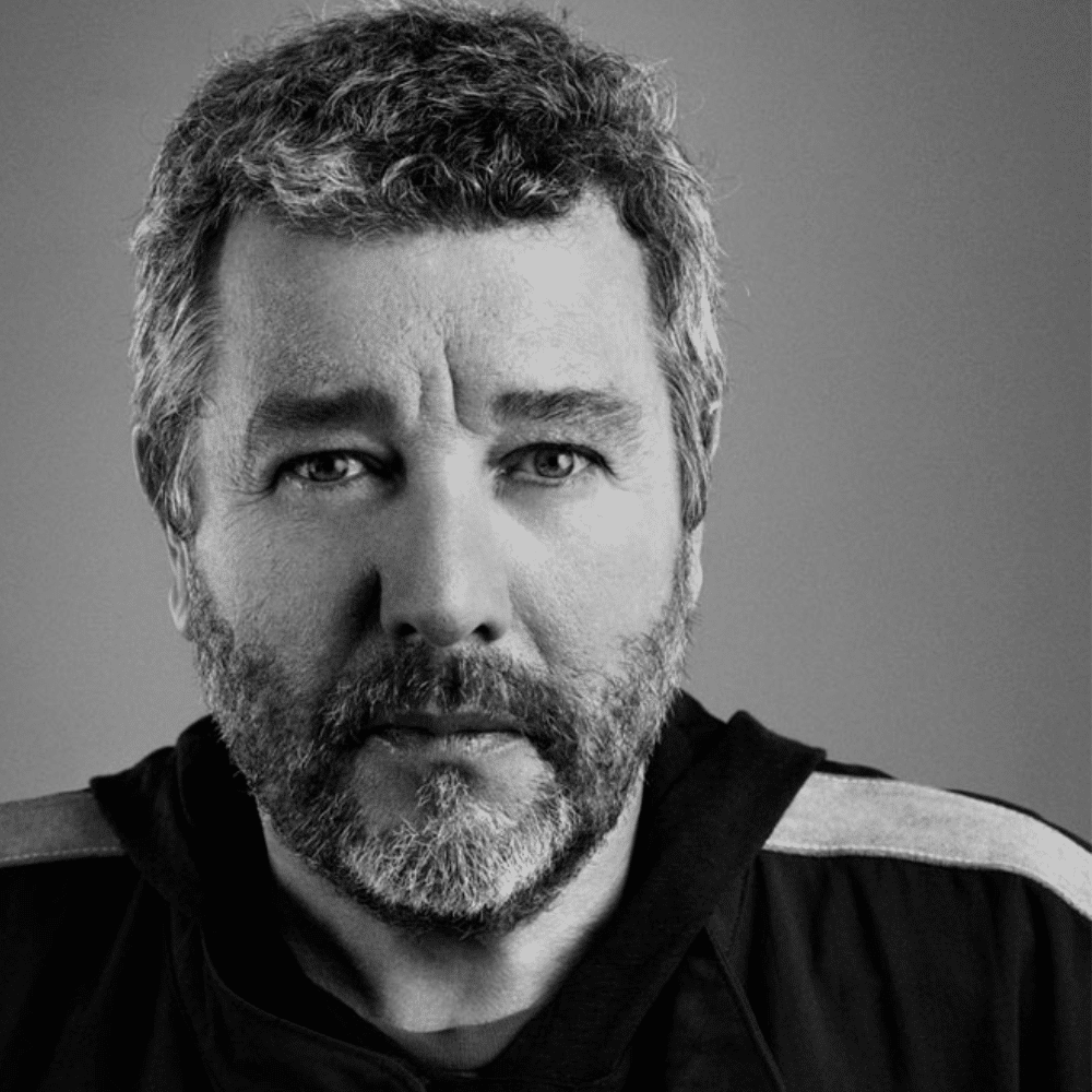 Black-and-white portrait of Philippe Starck with short curly hair wearing dark jacket against plain background.