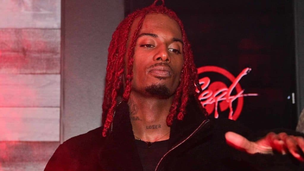 Rapper Playboi Carti with red dreadlocks wearing a black jacket under red ambient lighting.
