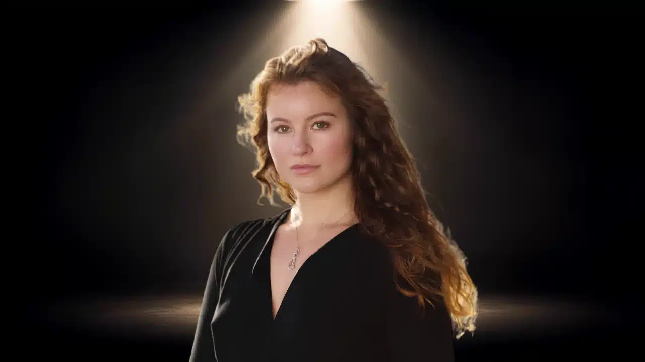 Portrait of Alexandra Andresen with wavy hair, wearing a black dress and necklace under light.