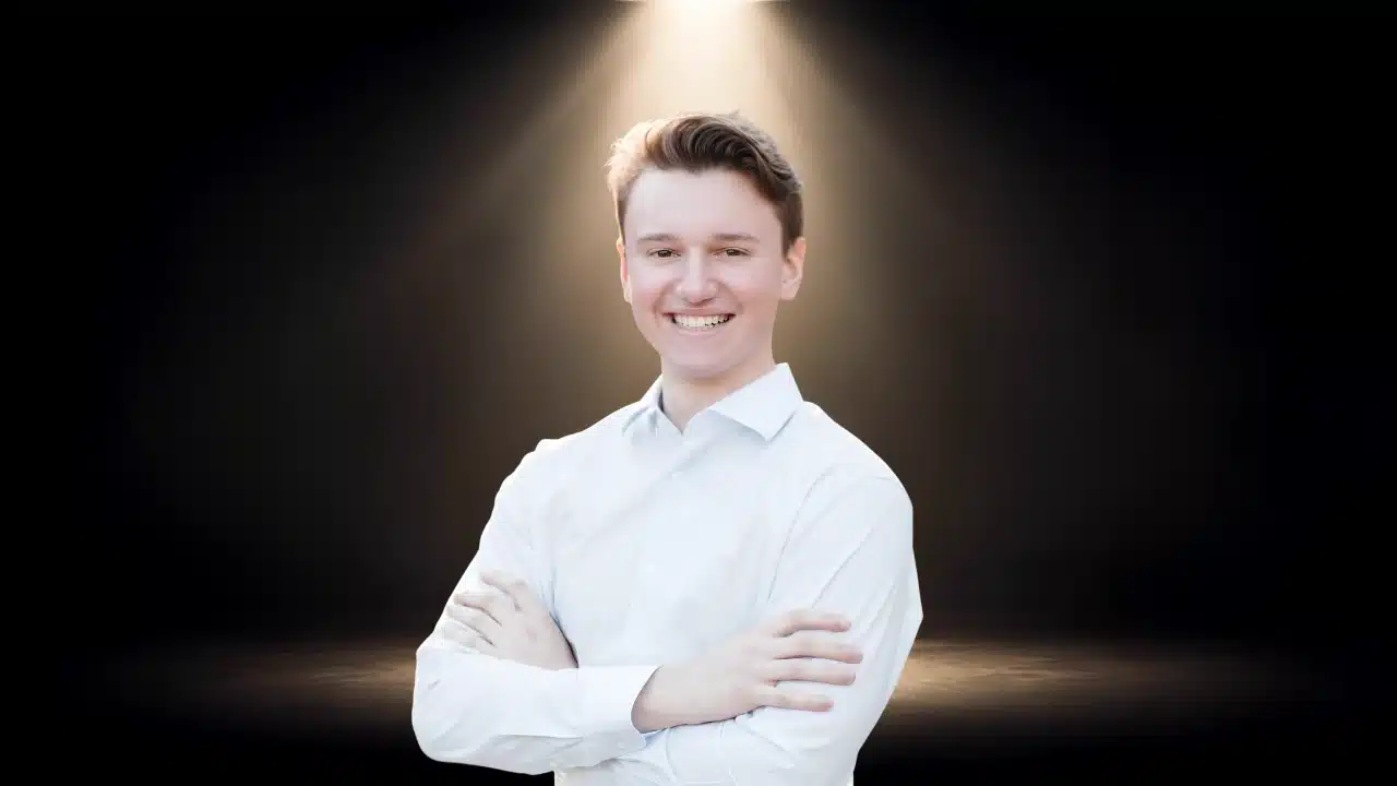 Portrait of Brendan Foody smiling with arms crossed, wearing a white shirt under a spotlight