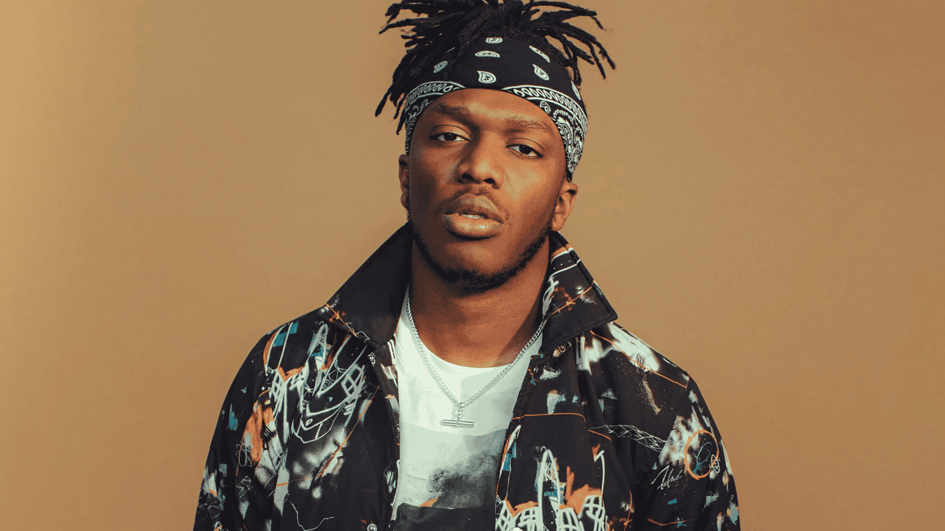 Portrait of YouTuber and musician KSI wearing a black paisley bandana and patterned jacket against a tan backdrop.