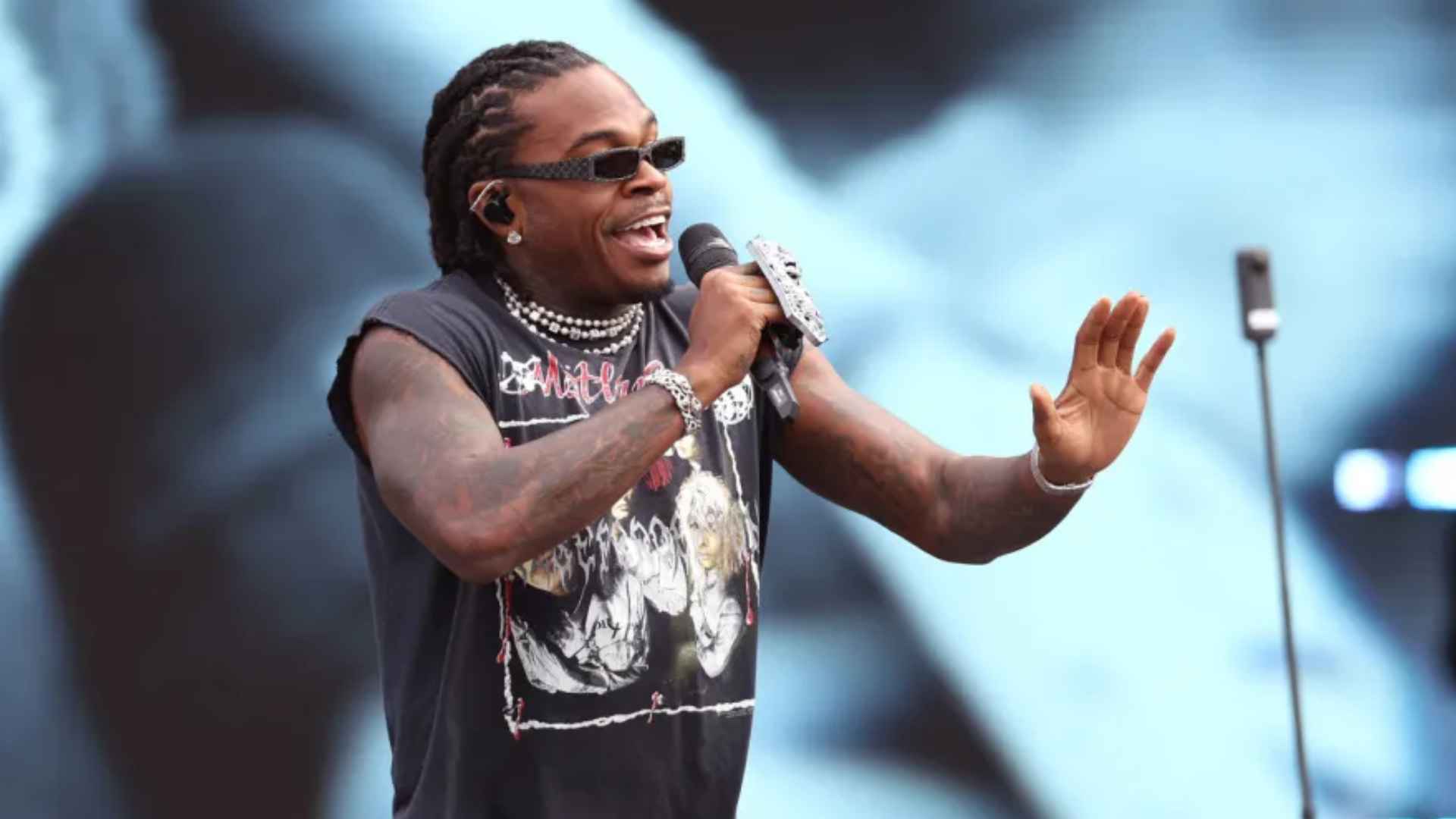 Rapper Gunna performing on stage with a microphone while wearing sunglasses and a black graphic shirt.