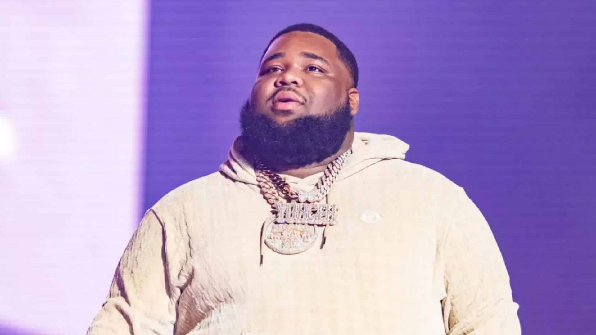 Rapper Rod Wave performing on stage, wearing a light-colored hoodie and diamond chains under purple concert lighting.