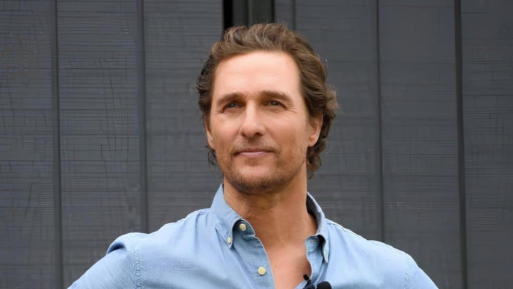 Matthew McConaughey wears a light blue button-down shirt and smiles while looking off-camera against a dark slatted backdrop