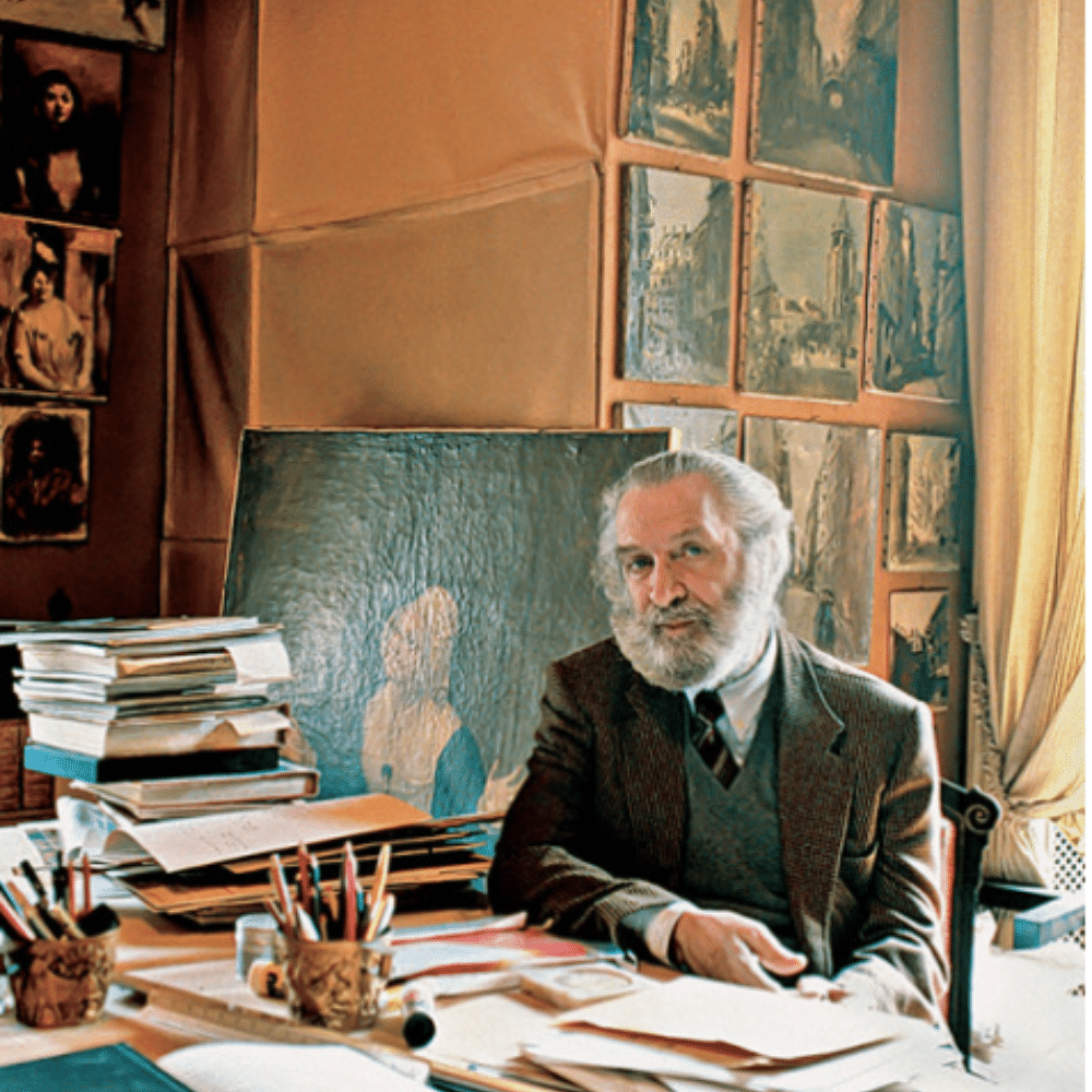 Renzo Mongiardino seated at cluttered desk in art studio surrounded by sketches, paintings, books, and papers.