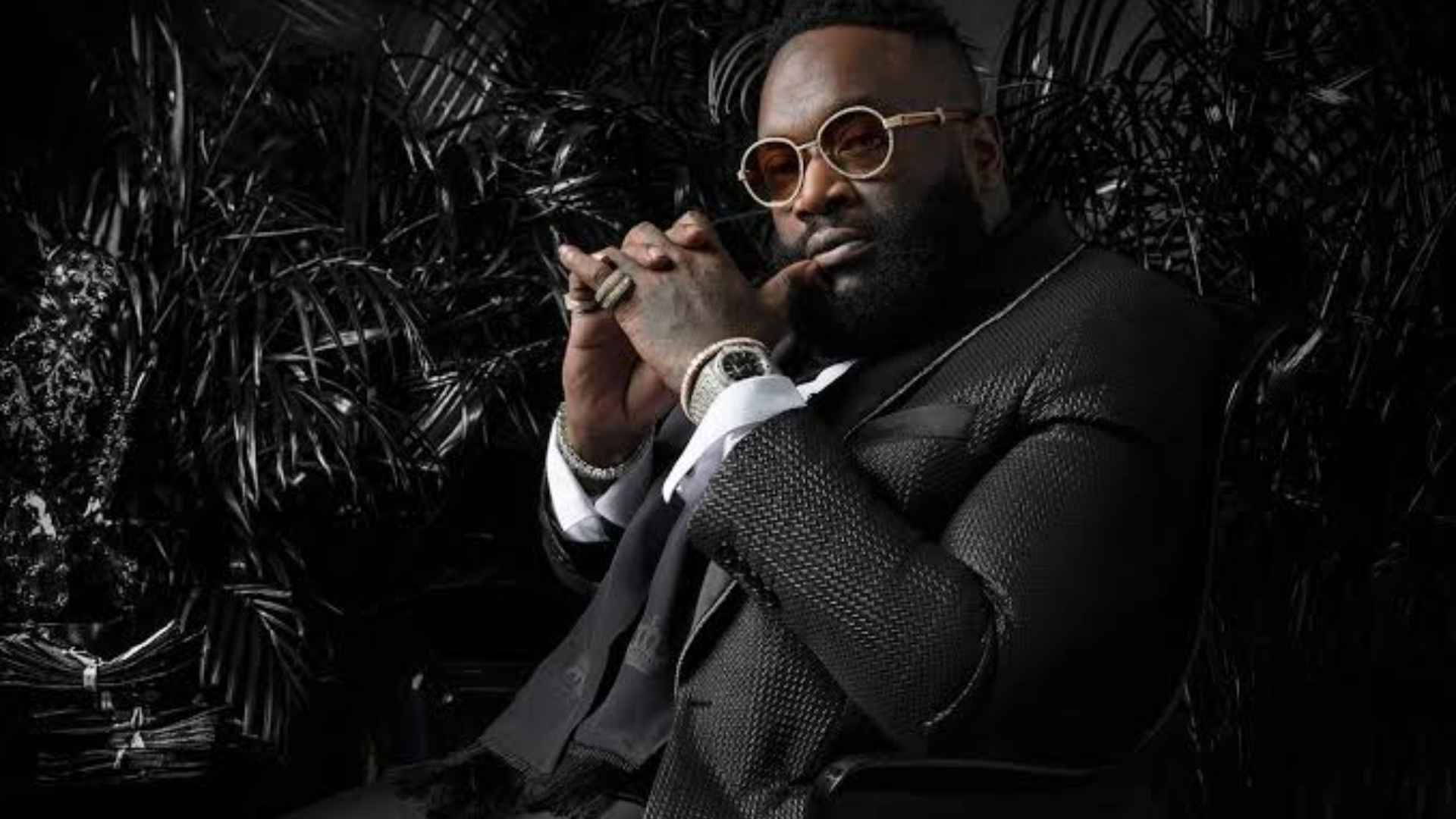 Rick Ross wearing sunglasses and a black suit while posing for a dark-themed portrait.