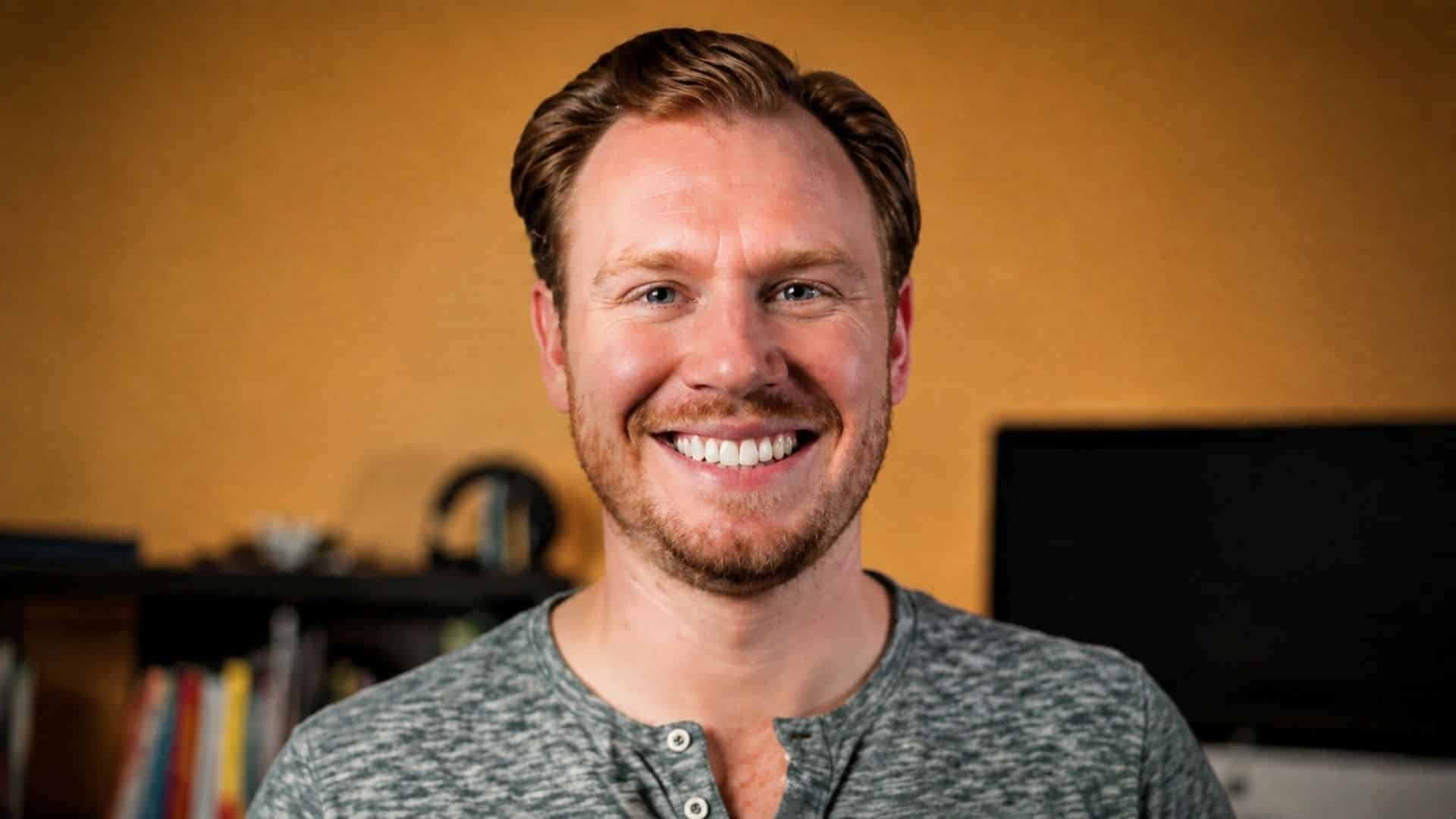 Riley Roberts smiling in a green gray shirt with warm indoor lighting and a softly blurred home office background