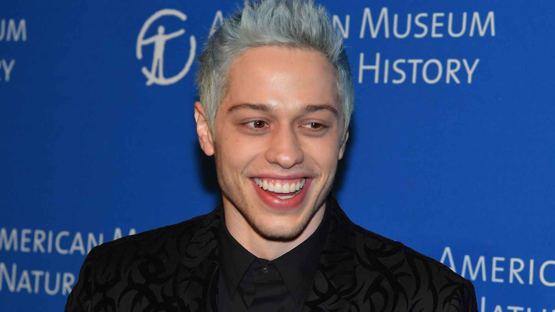 Pete with silver hair wearing a black suit at a formal event with a blue museum backdrop behind him