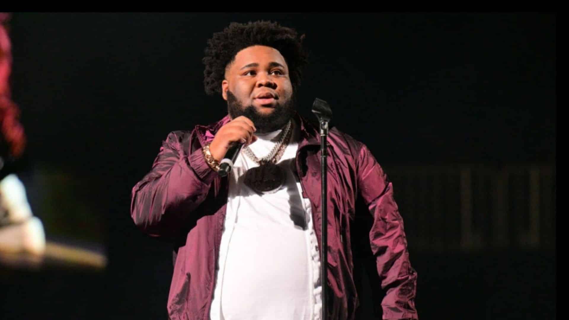 Rod Wave performing onstage, holding a microphone while wearing a burgundy jacket, white shirt, and layered chains.