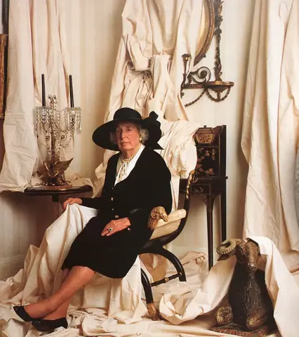  Sister Parish in black dress and wide-brim hat seated in elegant room with draped fabric, antique decor, and dog statue.