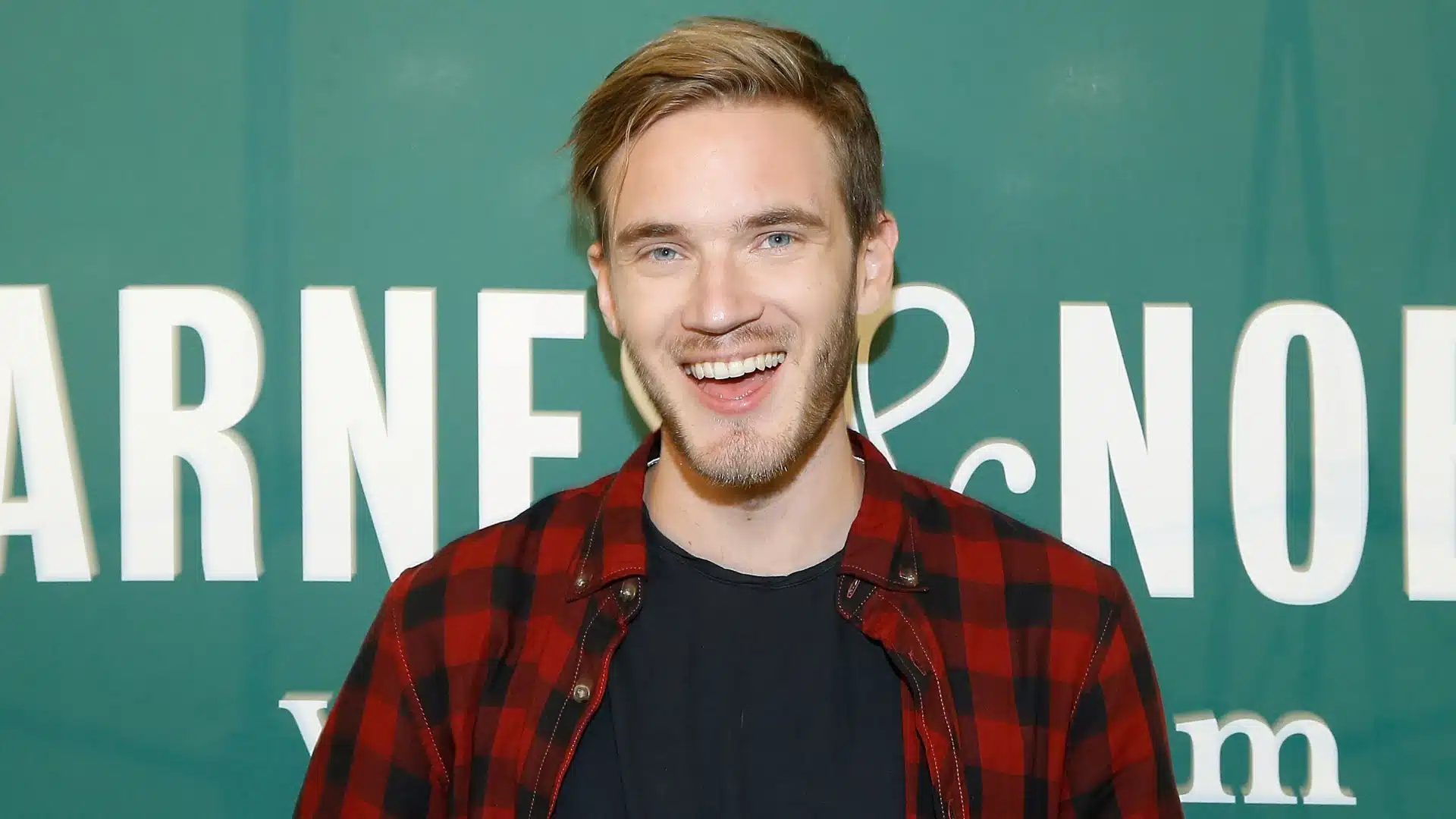 Smiling male YouTuber in a red plaid shirt posing at a public media event backdrop