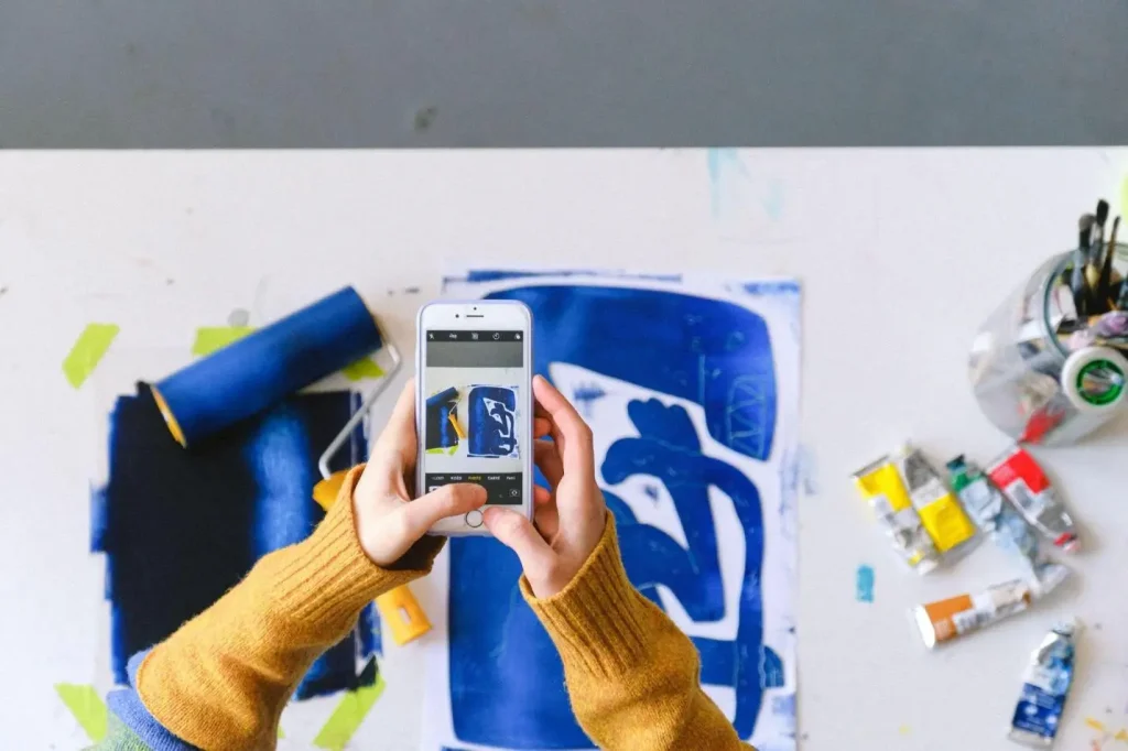 Person photographing blue artwork on a table with paint tubes and roller nearby