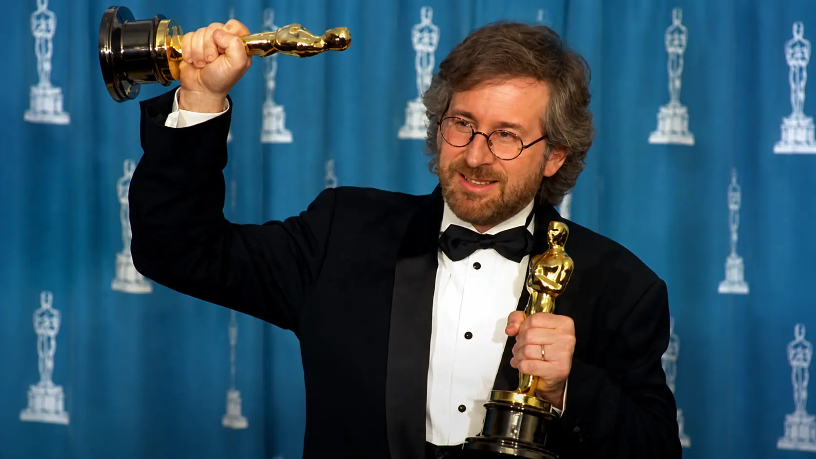 Steven Spielberg in a black tuxedo holding two golden awards at a ceremony with a blue backdrop and formal event setting area