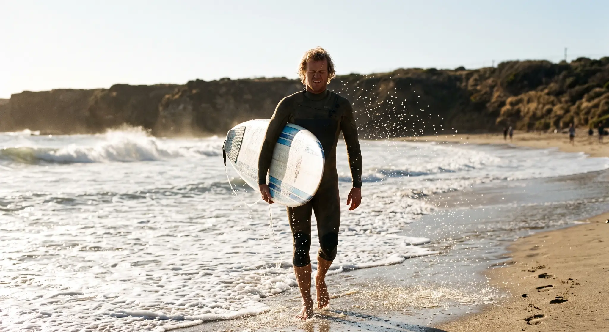 Surfer carrying a surfboard walks along sandy beach with waves in late afternoon sun