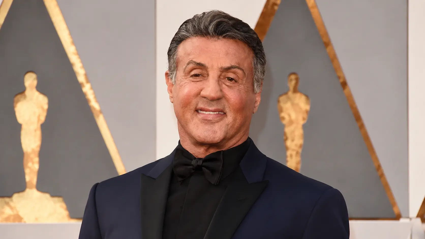 Sylvester Stallone wearing a navy tuxedo and black bow tie posing at an awards event with a formal backdrop and a warm smile