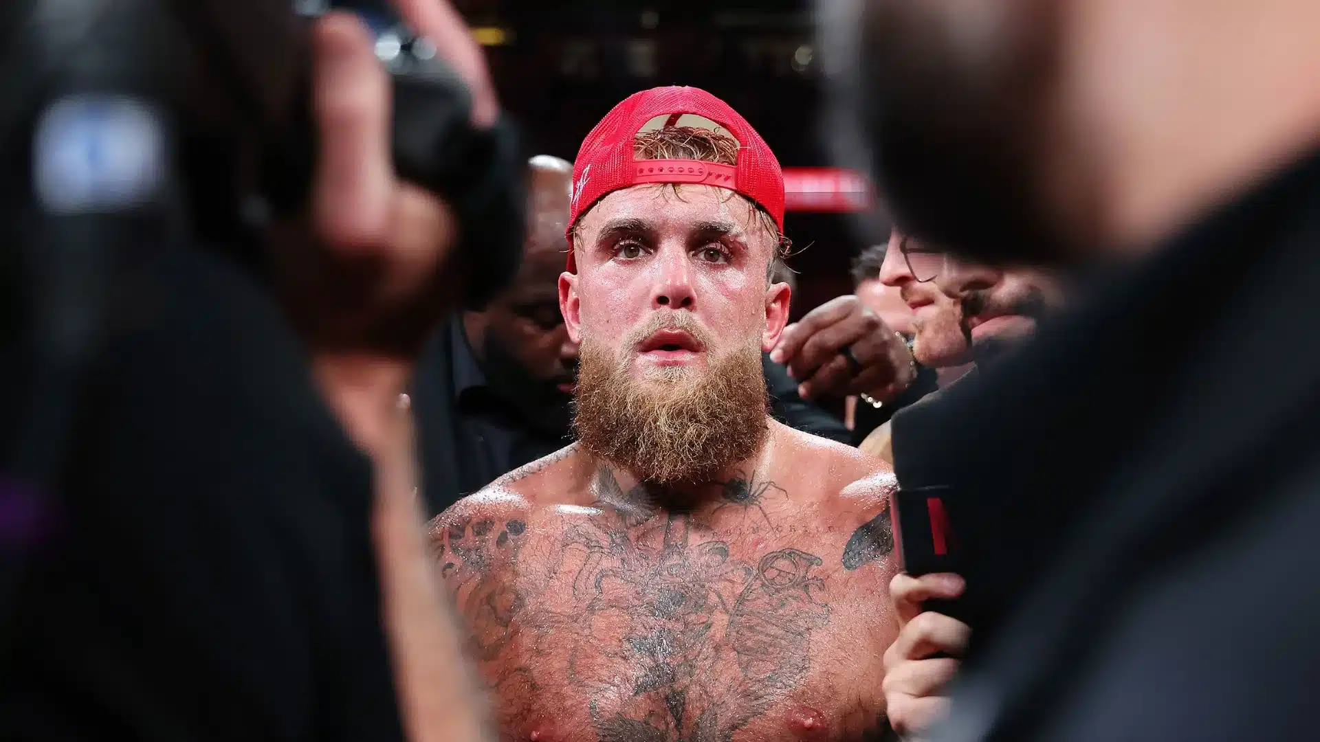 Tattooed boxer wearing a red cap while being interviewed inside a crowded boxing ring