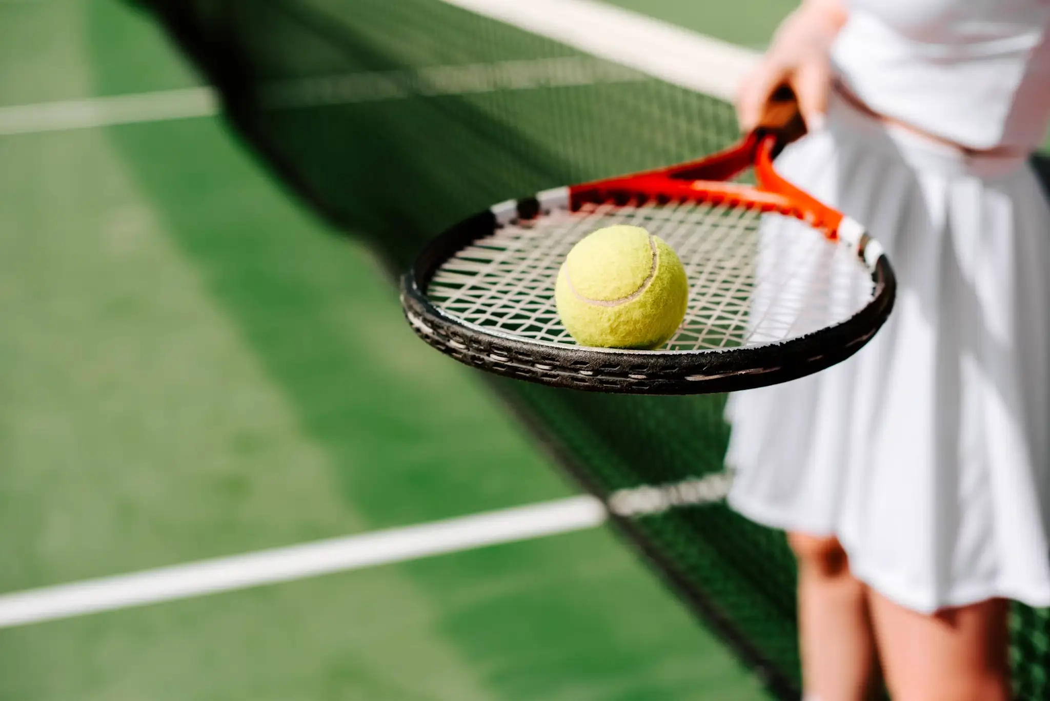 Tennis racket holding yellow tennis ball on outdoor court in bright sunlight