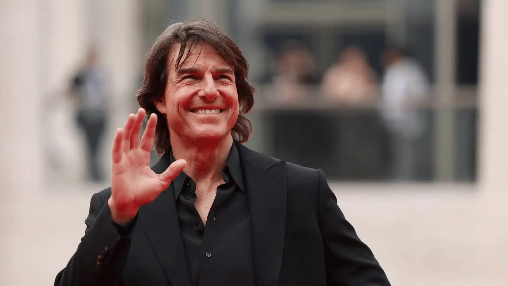 Tom Cruise smiling and waving outdoors in a black suit, with a blurred crowd and building in the background