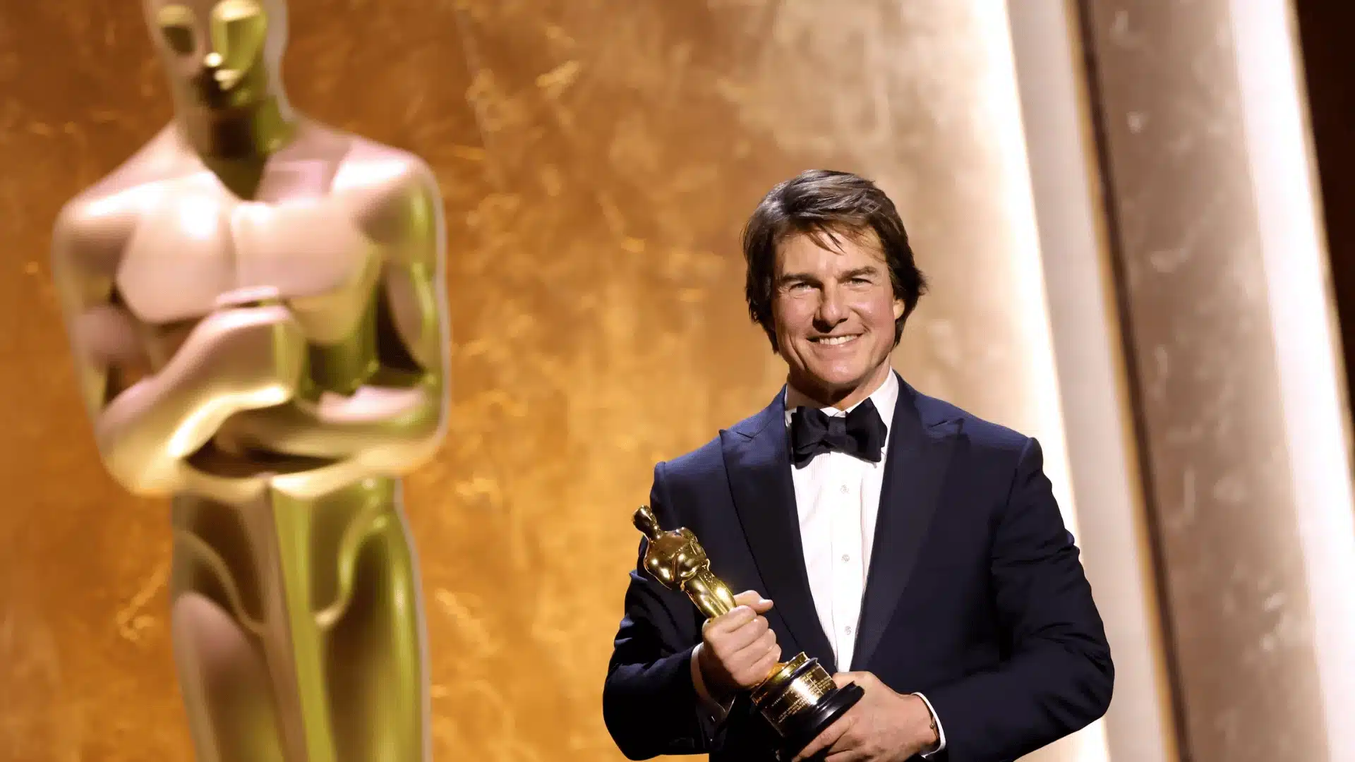 Tom Cruise smiling on stage while holding an Oscar statuette, with a large golden Oscar statue in the background