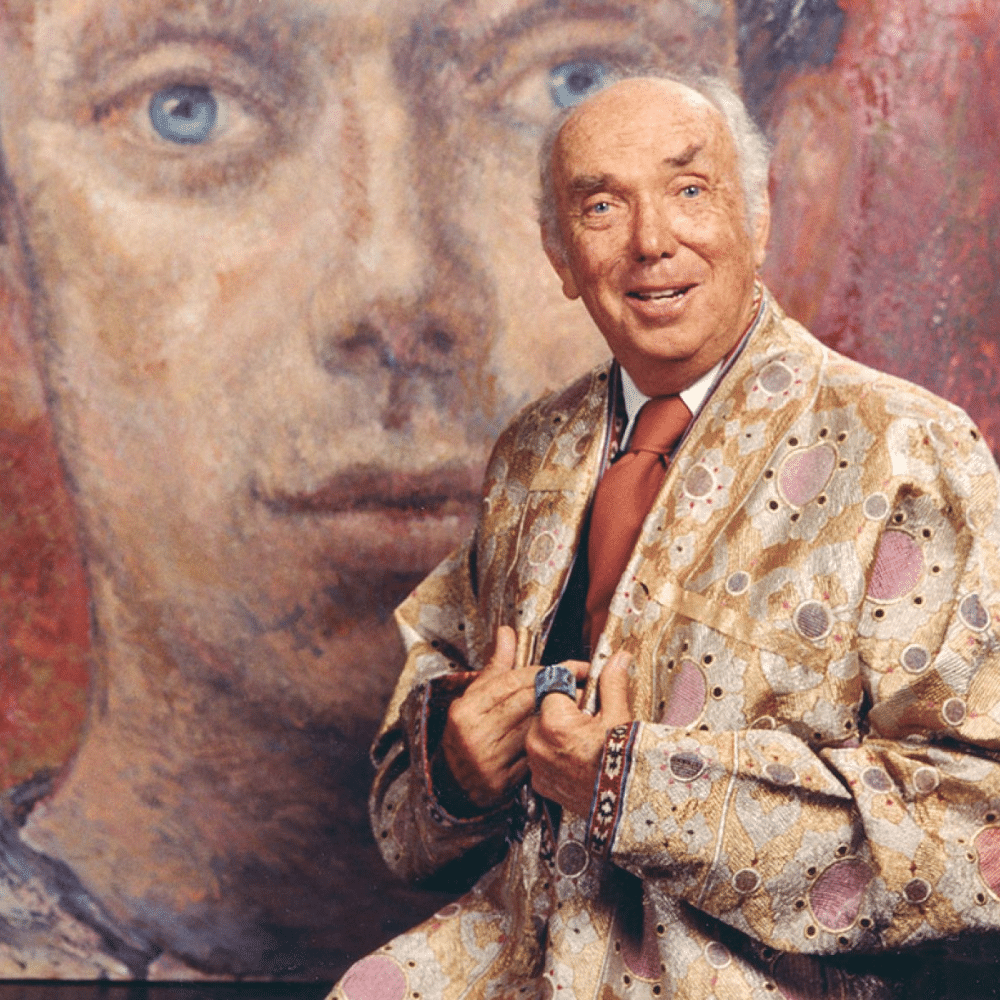 Tony Duquette in patterned jacket posing before large painted portrait with blue eyes in colorful art studio setting.